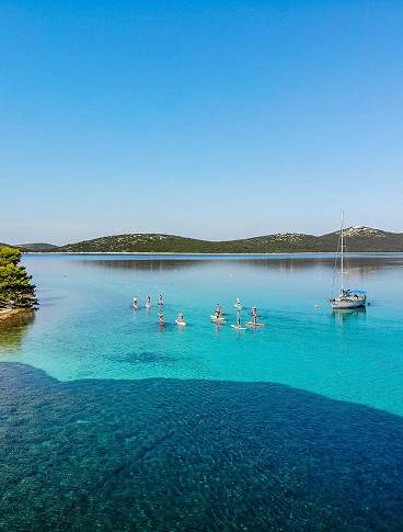 People paddleboarding on a clear turquoise lake with a moored boat and distant hills under a blue sky