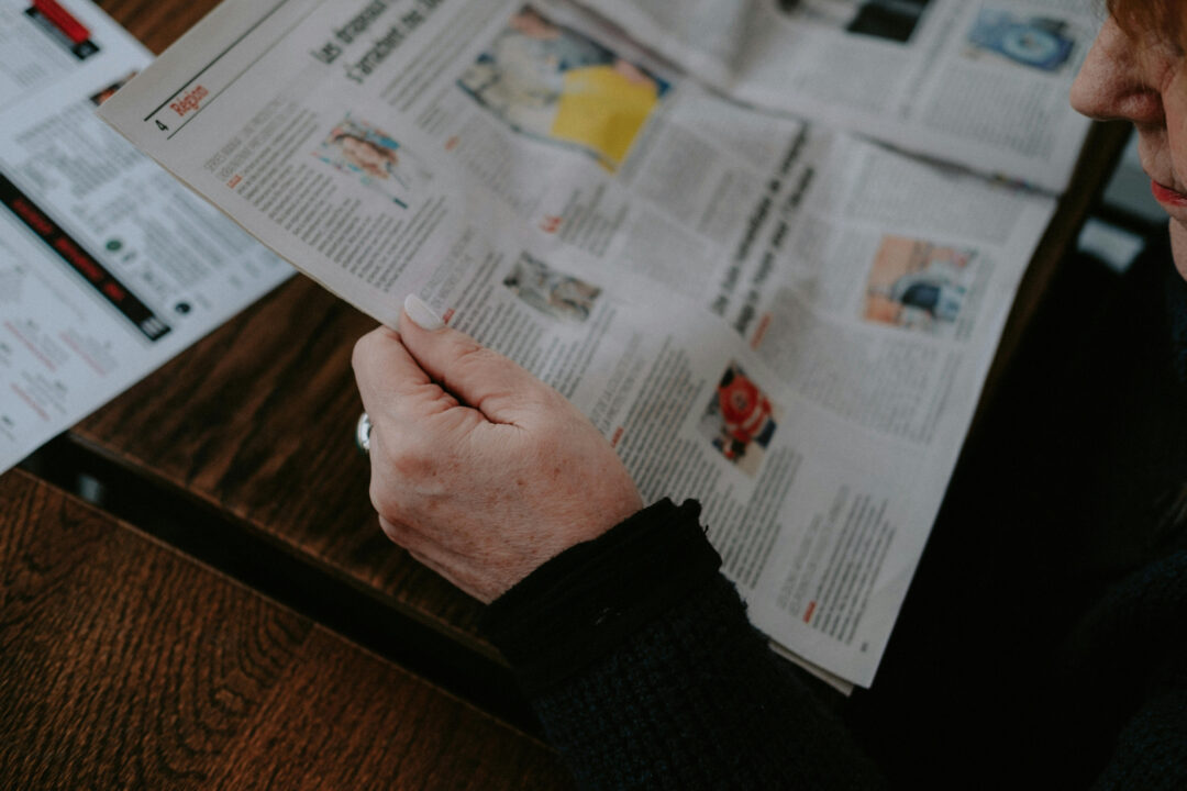 Hands holding an open newspaper with printed articles and photos on a table