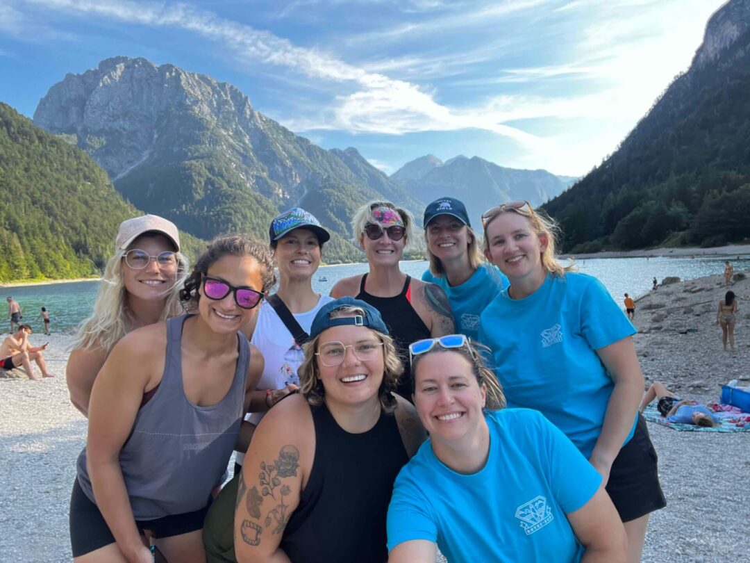 Women posing on a lakeside beach with mountains behind them, with others swimming in the water