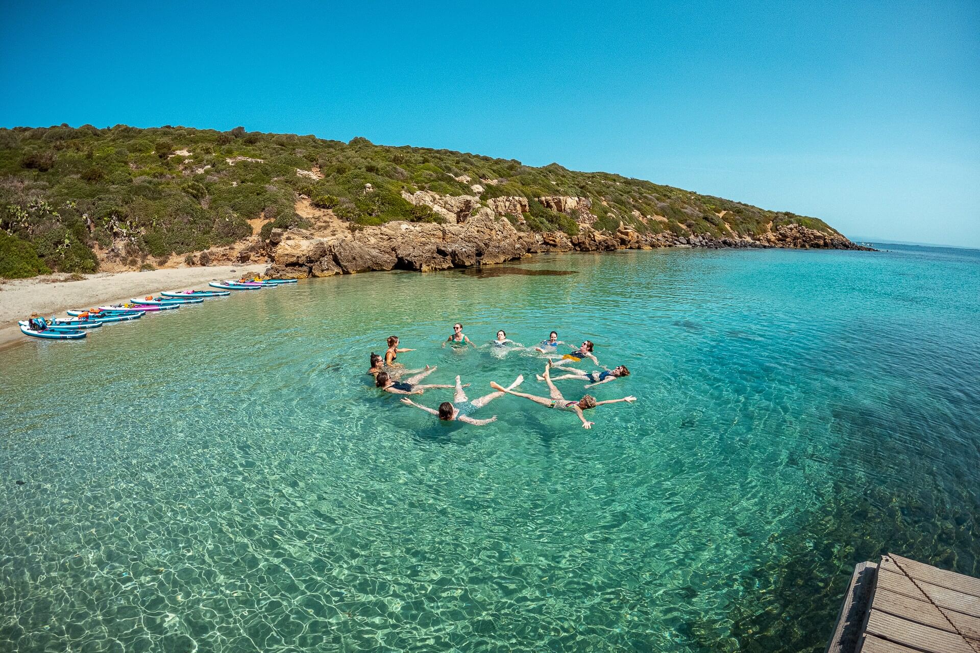 Women swimming in clear coastal water with paddleboards moored near a rocky shoreline