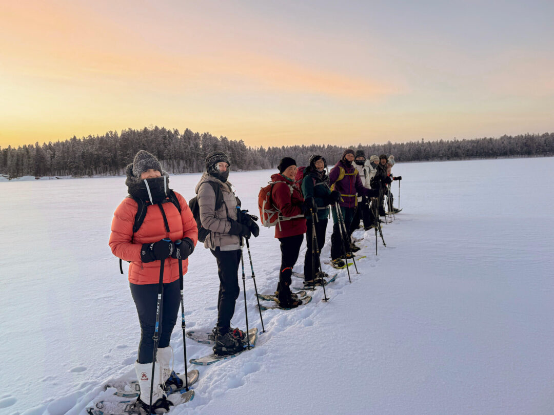 Women skiing in snowy Finland at sunrise, a group walking on skis with poles across a frozen lake