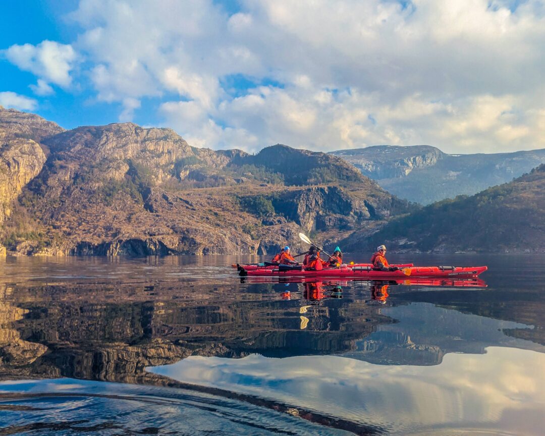 Women paddling red kayaks on a calm lake beside rocky mountains under a blue sky