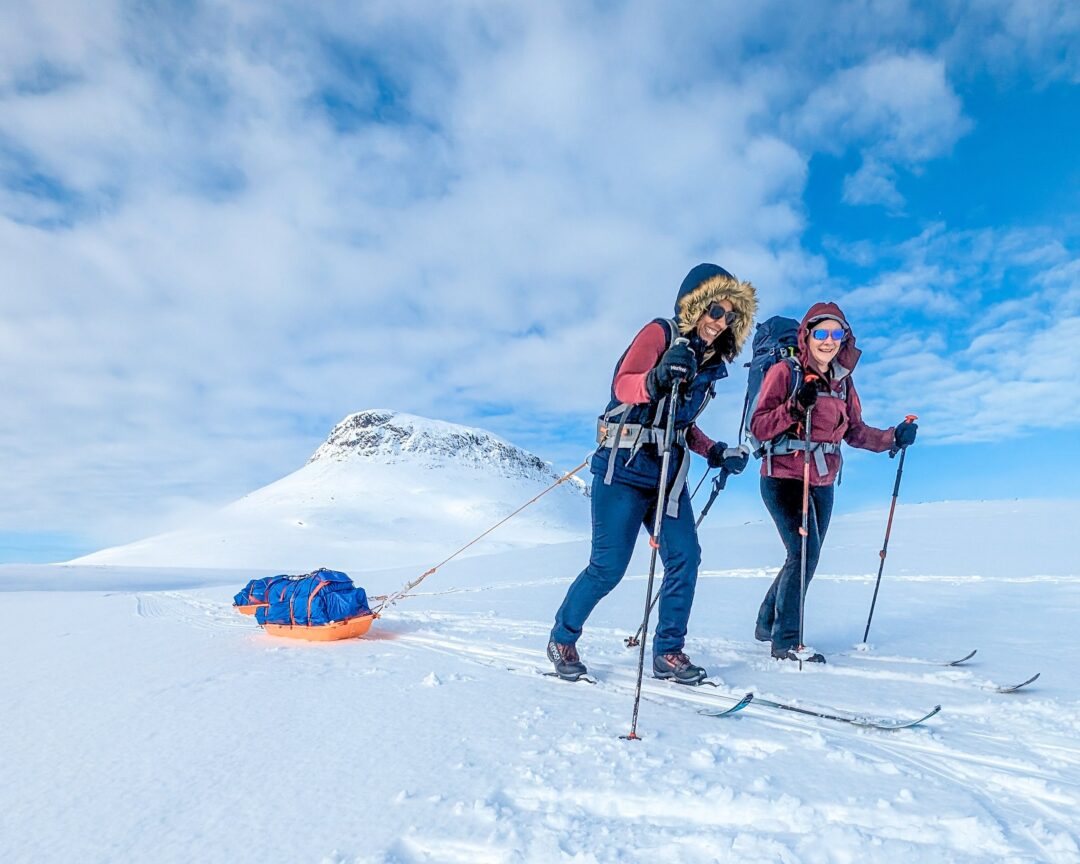 Two women ski touring on a snowy mountain with skis and poles, with a sled in tow.