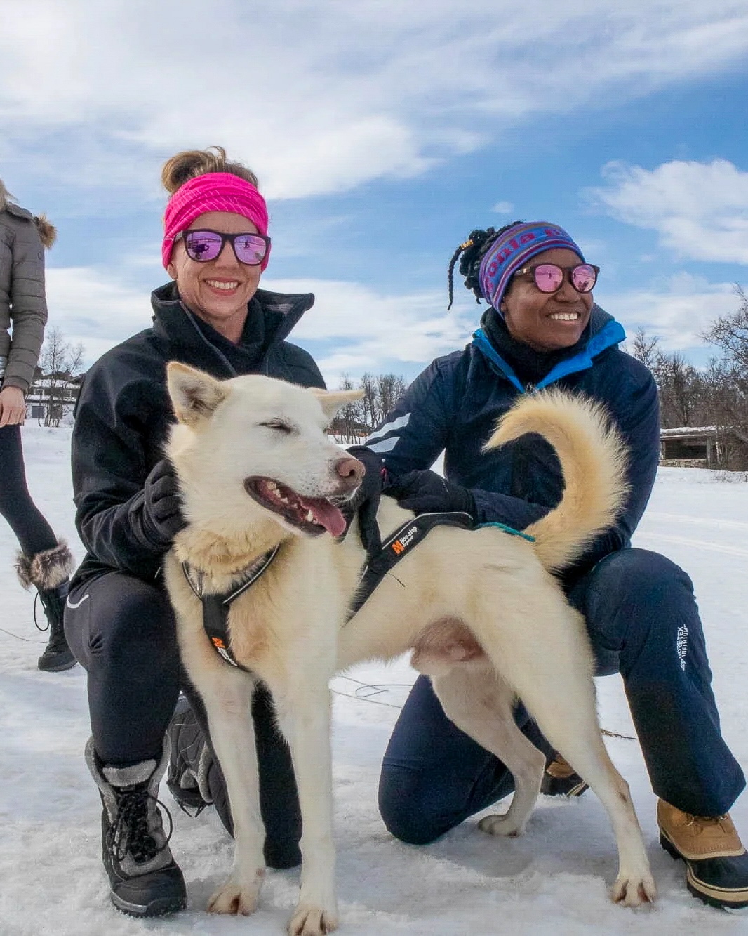 Two women kneeling in snow with husky dogs outdoors, wearing winter hats and sunglasses