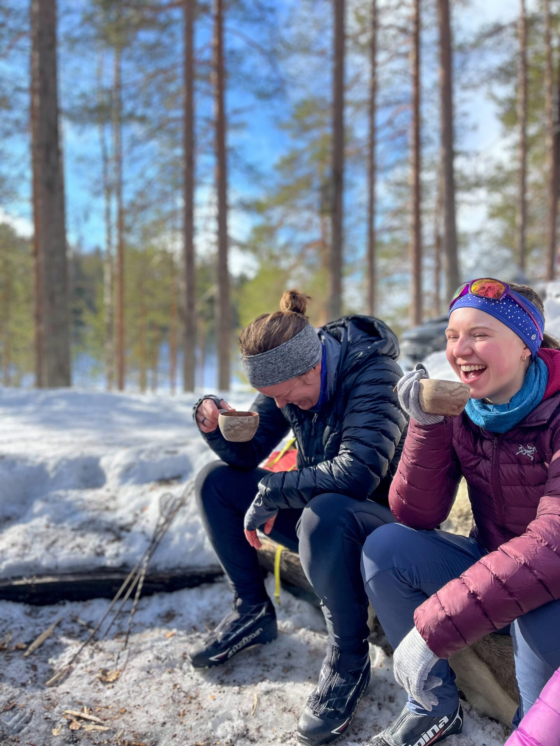 Women in winter jackets and headbands laughing while drinking hot drinks in a snowy forest