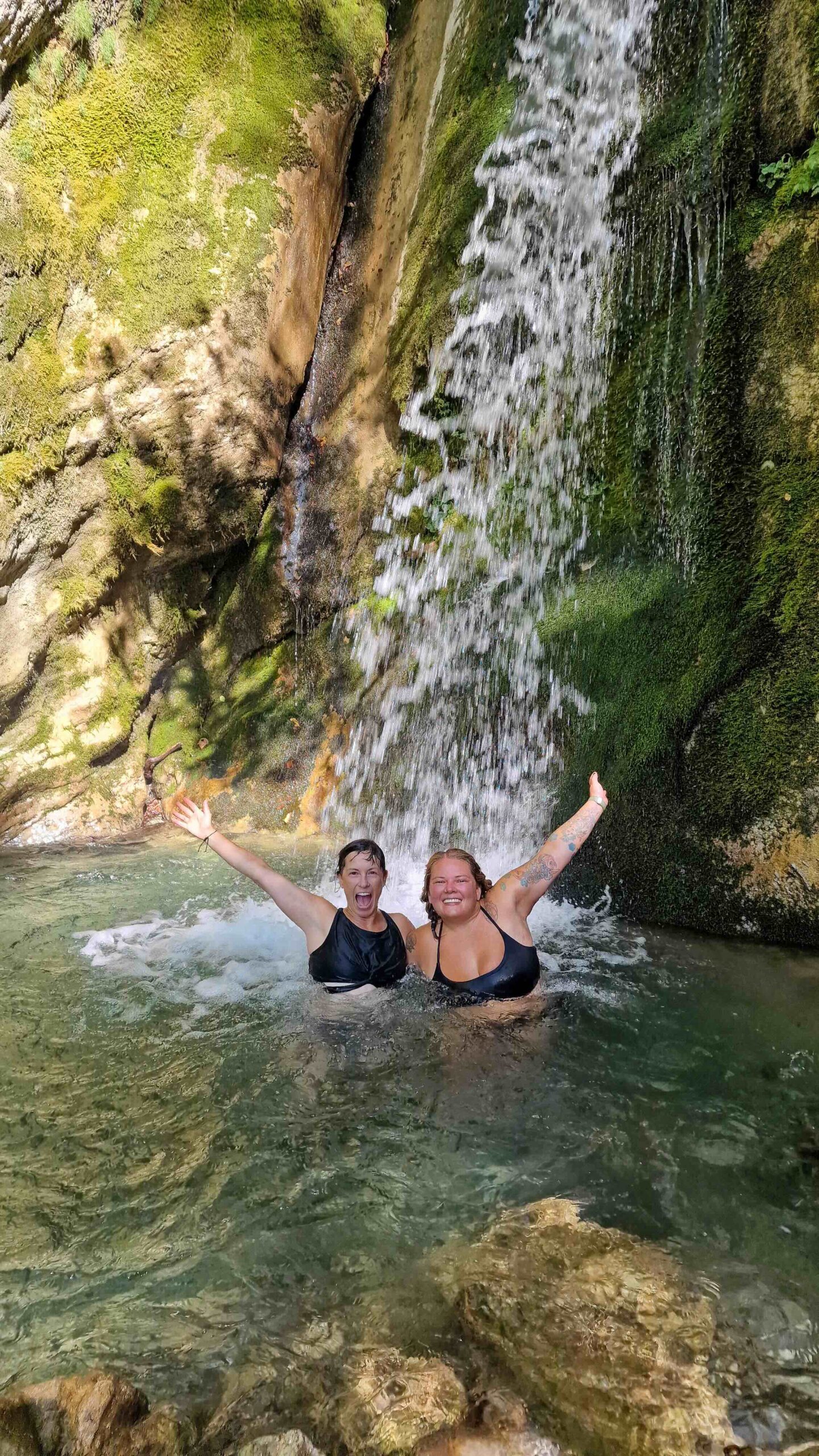 Two women in a shallow pool beneath a waterfall in a rocky gorge, arms raised.