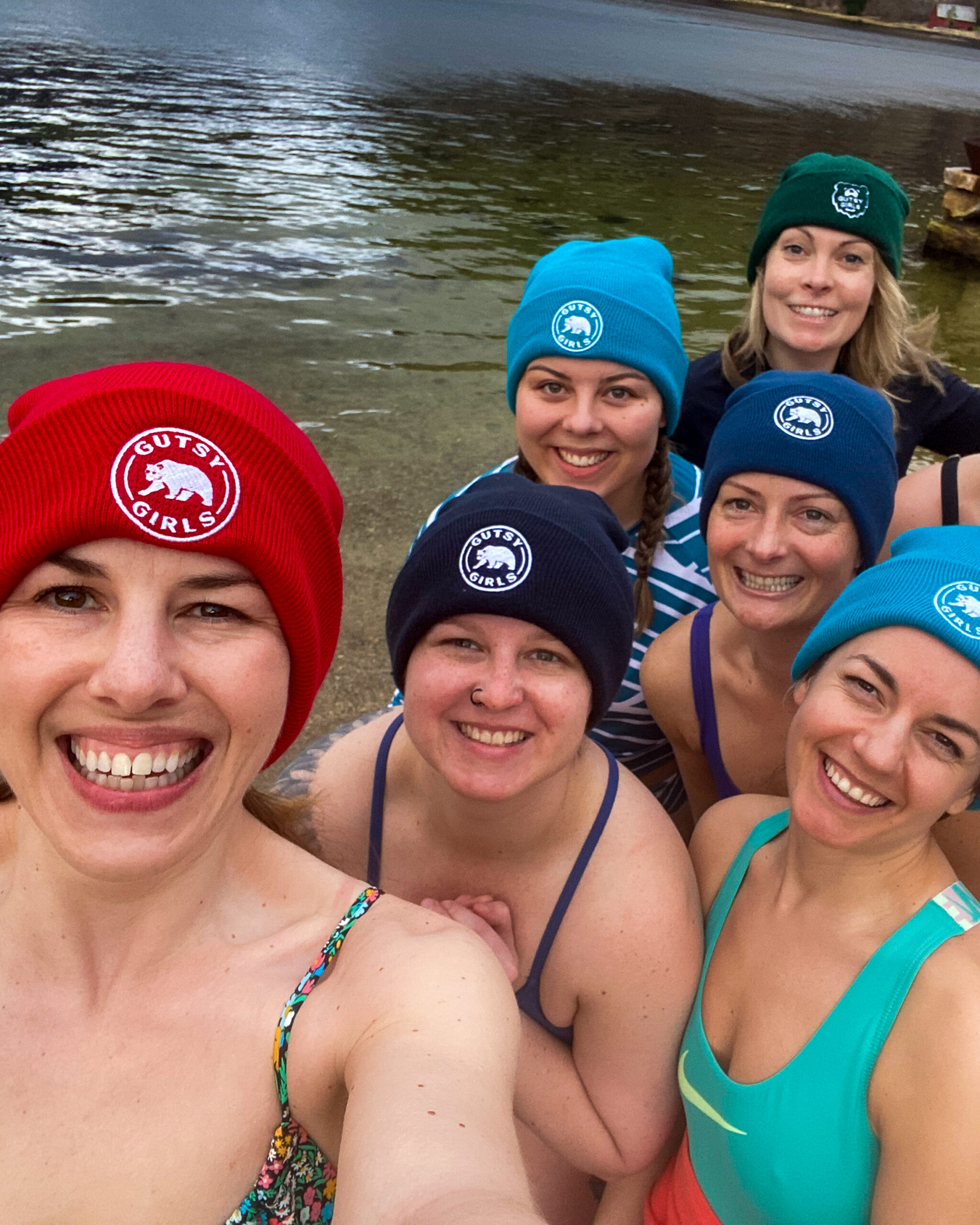 Smiling women in swimsuits wearing Gutsy Girls beanies in open water by the shore