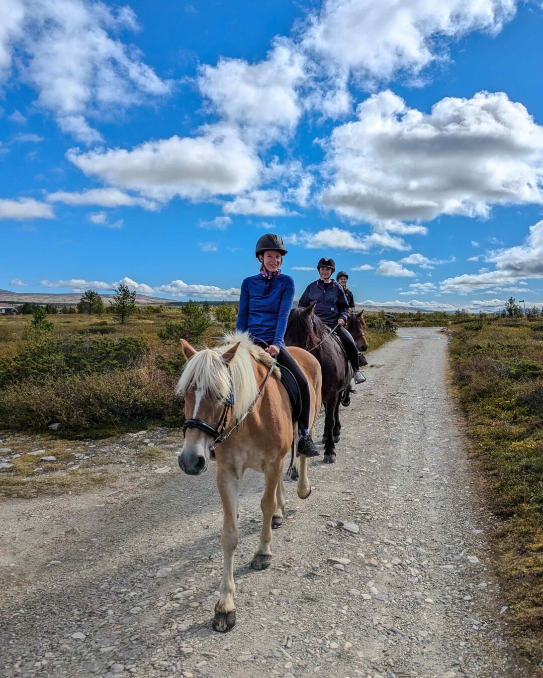 Women riding horses along a gravel path in open countryside under a cloudy blue sky