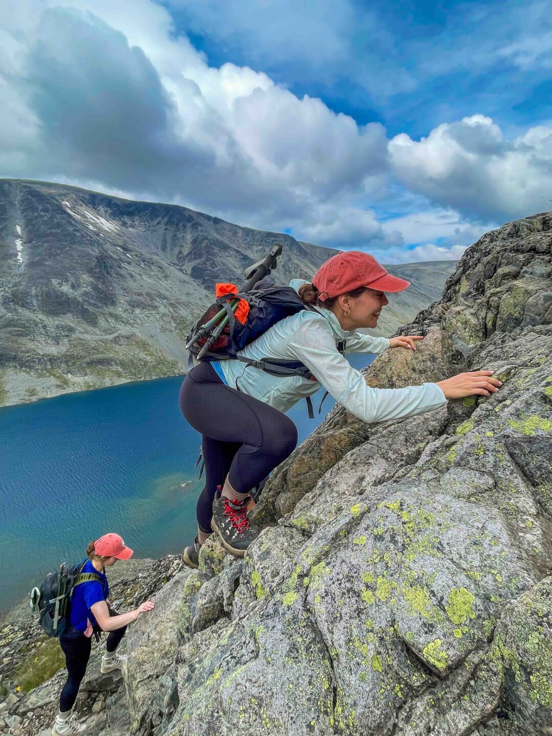 Hiking: women on a rocky mountainside with backpacks, overlooking a lake under cloudy skies