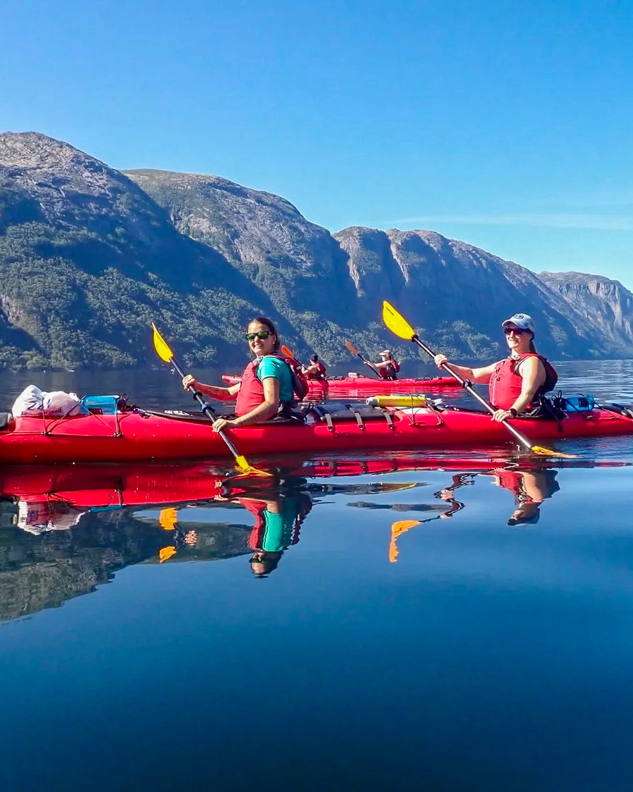 Two women kayaking in a fjord, red kayaks and yellow paddles against mountain scenery