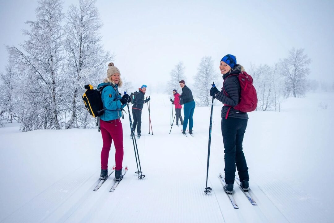 Women cross-country skiing on a snow-covered trail through a winter forest, wearing ski poles and boots.
