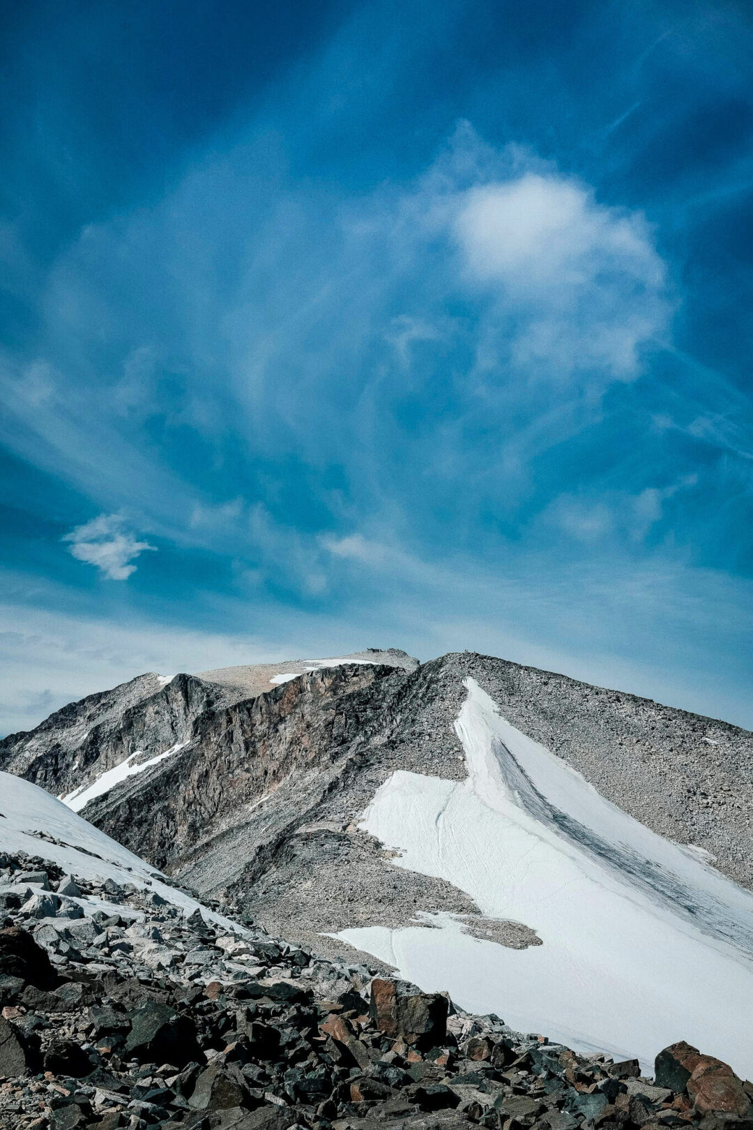 Snowy mountain ridge in Norway with rocky foreground and a narrow glacier under a blue, cloudy sky