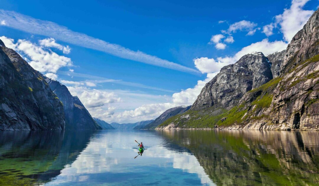 Woman kayaking on a calm fjord with steep mountains and a clear blue sky.