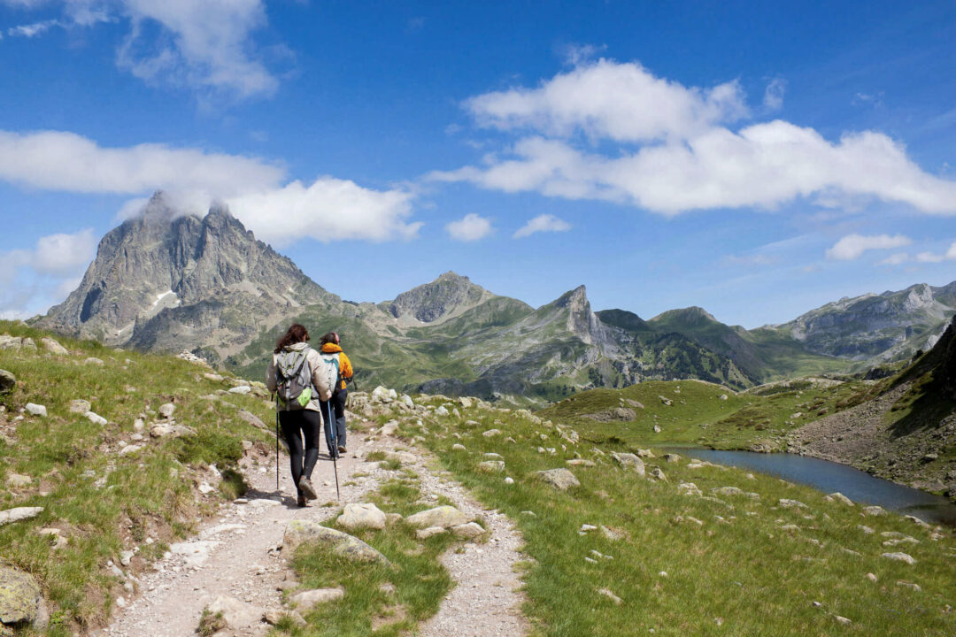 Women hiking on a mountain trail overlooking a lake in an alpine landscape with peaks and clouds