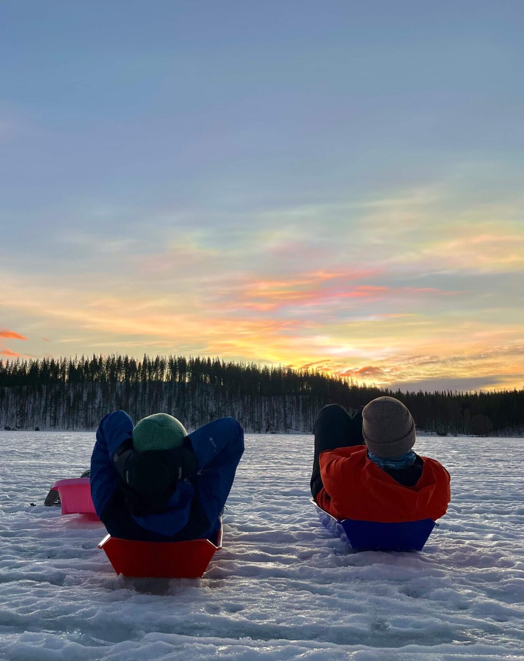 Two women in winter jackets sit on sleds on a frozen lake at sunset, with a snowy forest behind