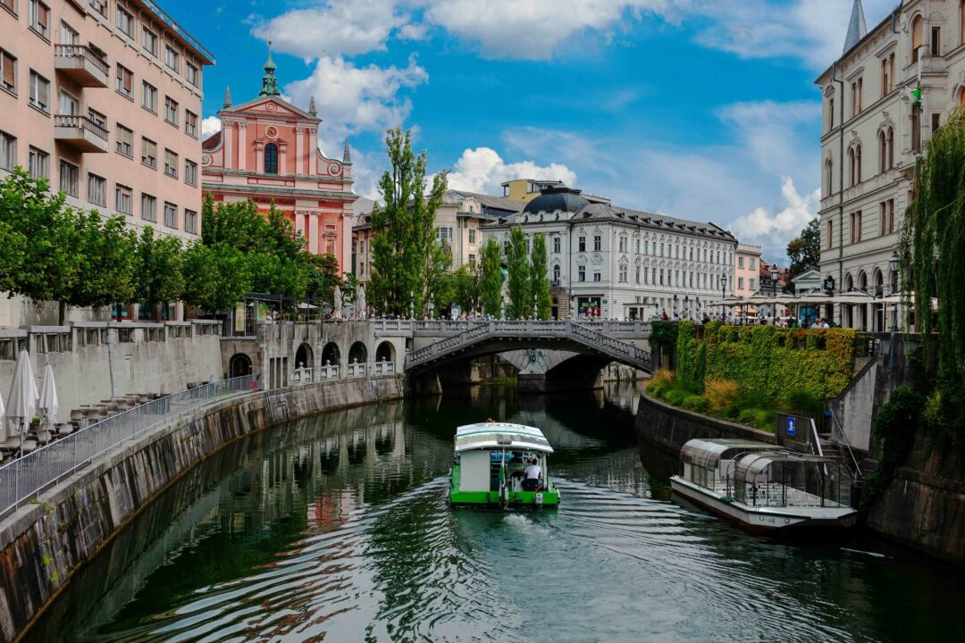 Boat on the Ljubljanica River in Ljubljana, with a stone bridge and riverside buildings under a blue sky