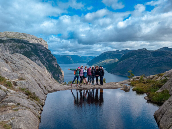 Women hikers pose on a rocky ledge overlooking a fjord under a cloudy sky