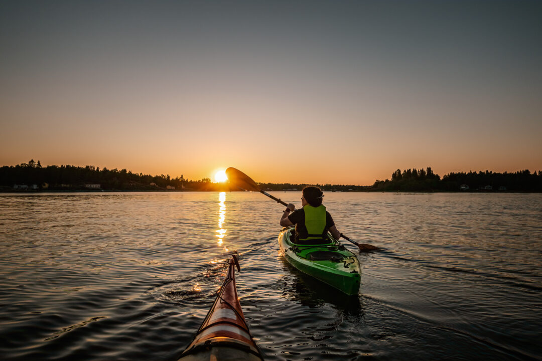 Women kayaking on a calm lake at sunset, with the sun low on the horizon and a second boat visible in front
