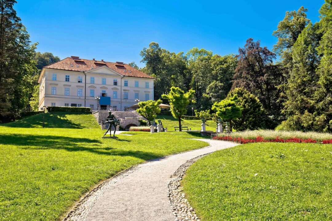 Landscaped park grounds with a historic mansion, statues and trimmed flowerbeds under a blue sky