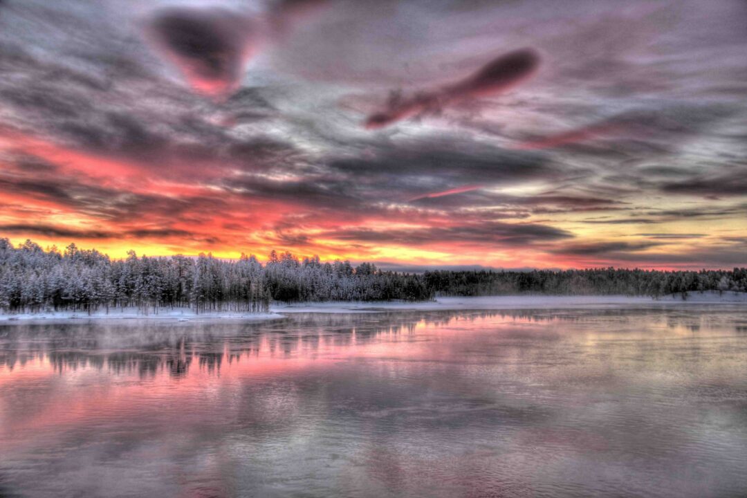 Aurora borealis over a snowy Arctic lake at sunset, with trees and still water reflections