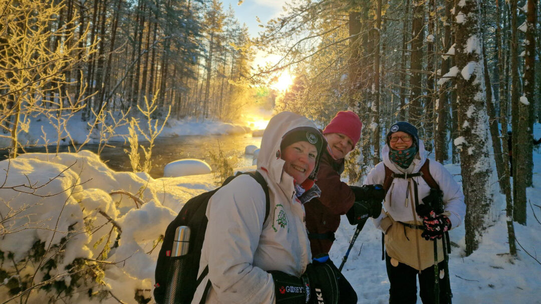 Women snowshoeing in a snowy pine forest at sunrise in Finland, smiling with trekking poles