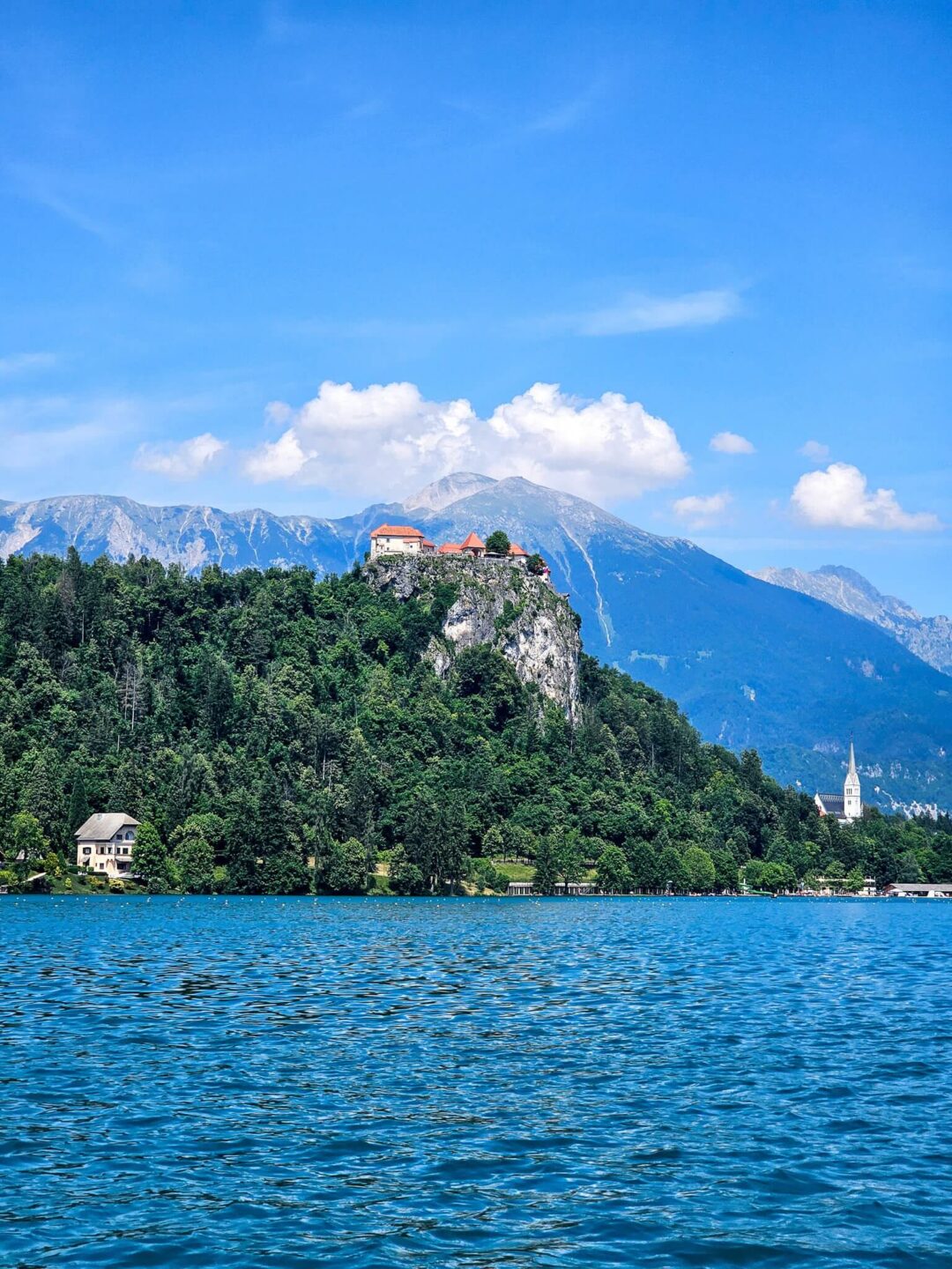 Alpine lake view with forested hills and a fortress on a cliff under a blue sky