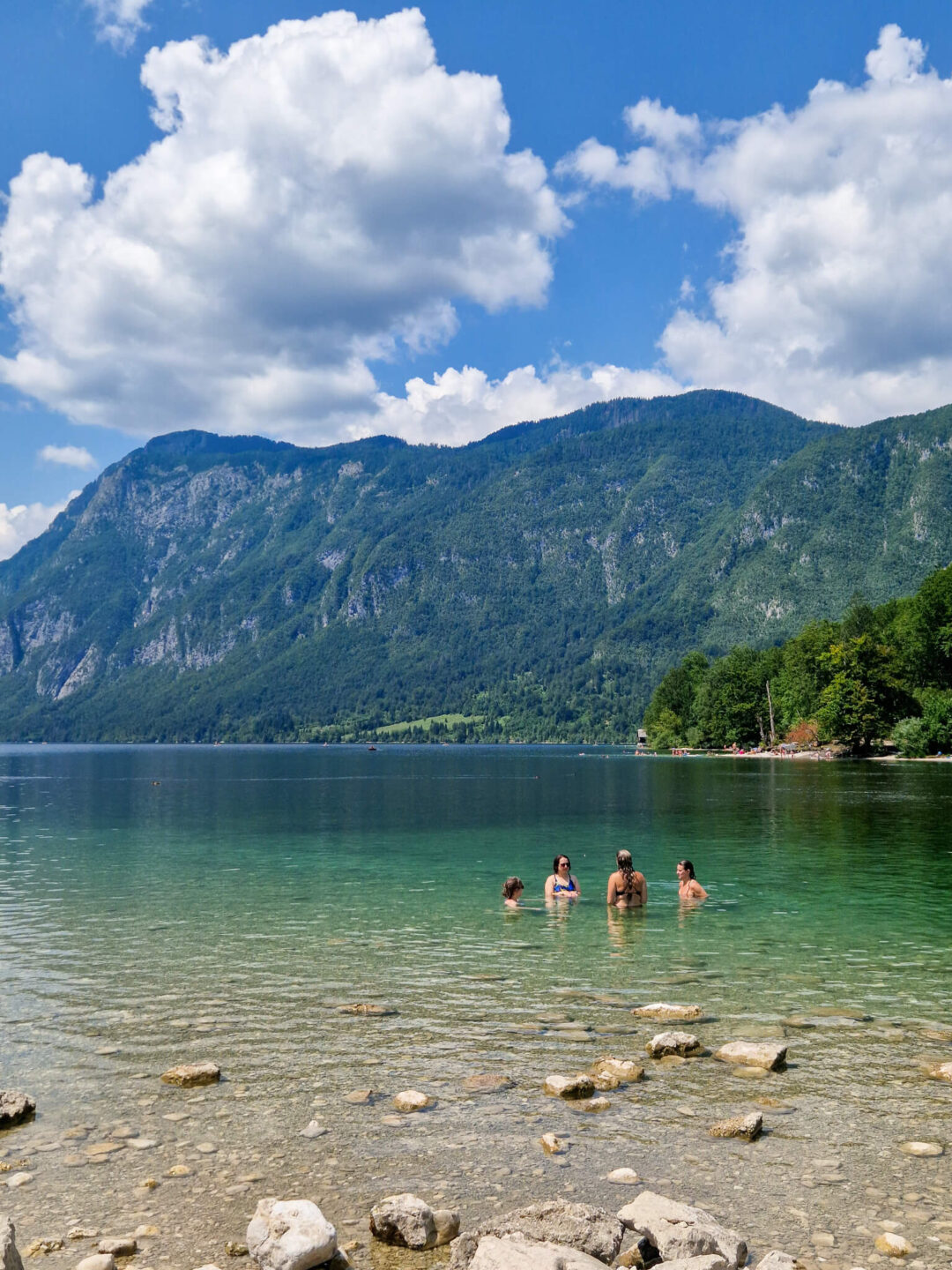 Women wading in a calm lake with forested mountains and blue sky