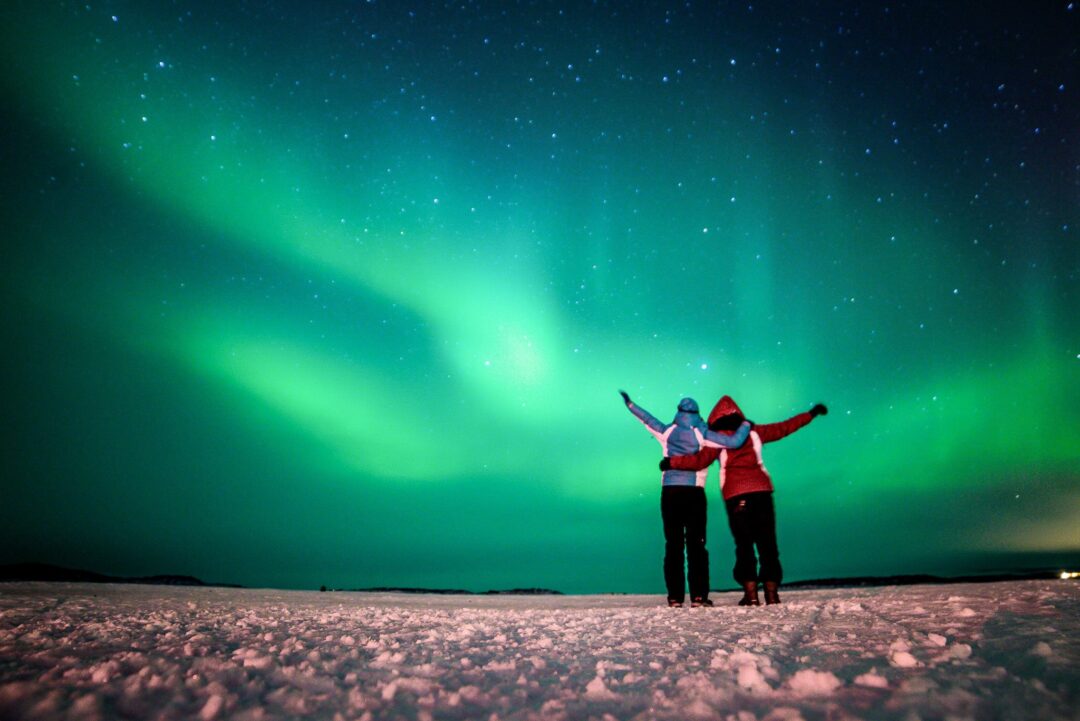 Two women standing on snowy ground beneath the northern lights (aurora) at night