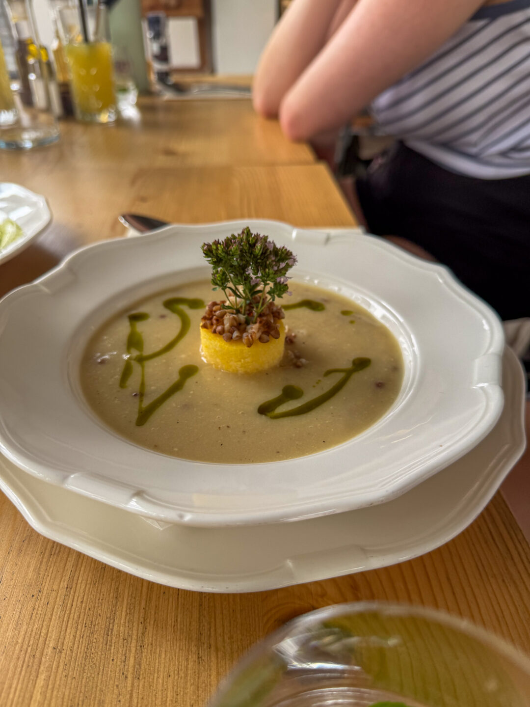 Plated soup with herb garnish served at a table, with a woman’s arm in the background