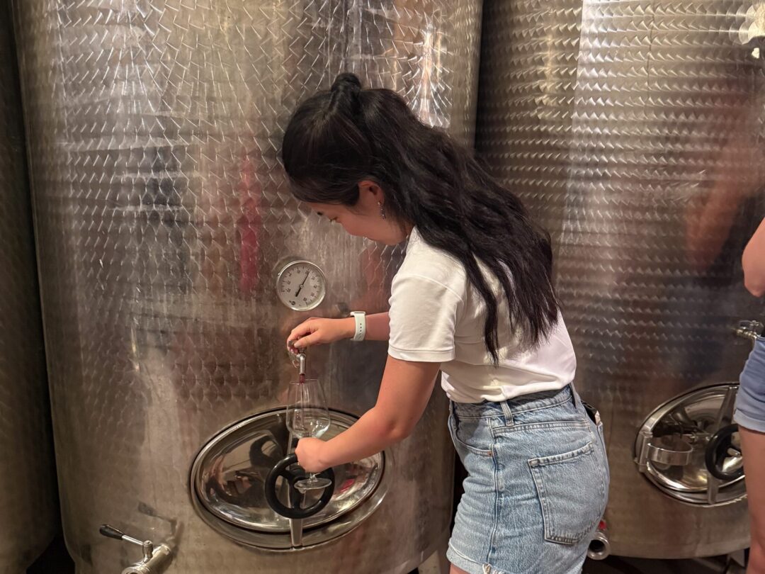 Woman operating a winery tap to pour wine from large stainless-steel tanks