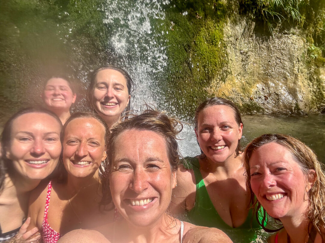 Women smiling in a river near a waterfall, cooling off together during an outdoor adventure holiday