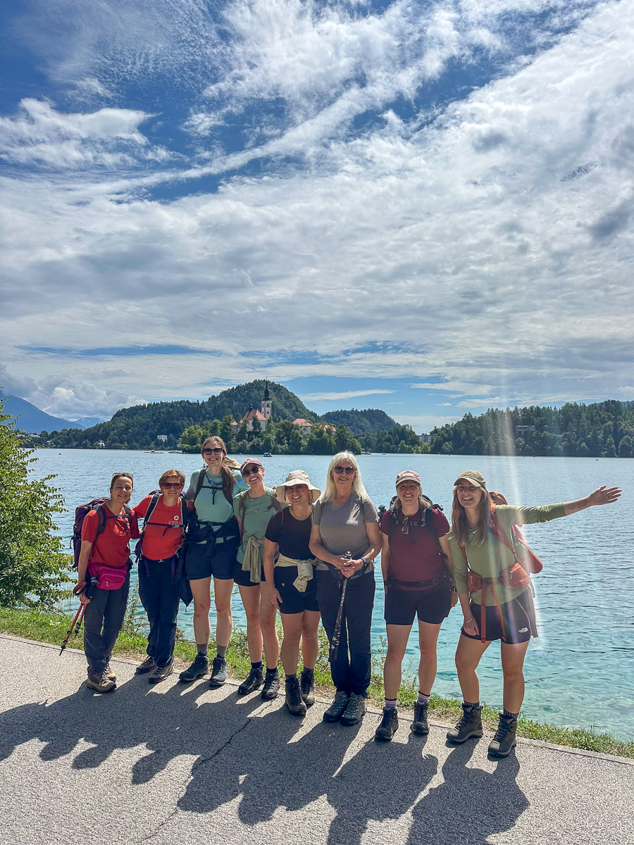 Women hiking group posing by a lake with a hillside castle in Slovenia under blue skies