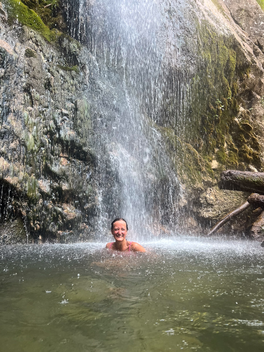 Woman in a mountain waterfall pool, smiling as water cascades down rock face