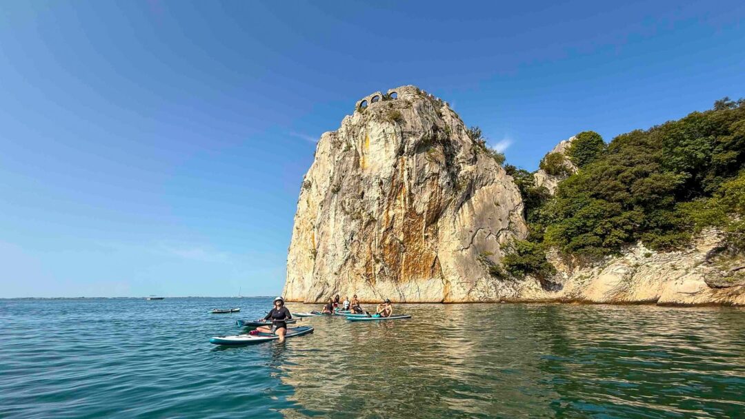 Woman paddleboarding on calm water beside a rocky coastal cliff and beach.