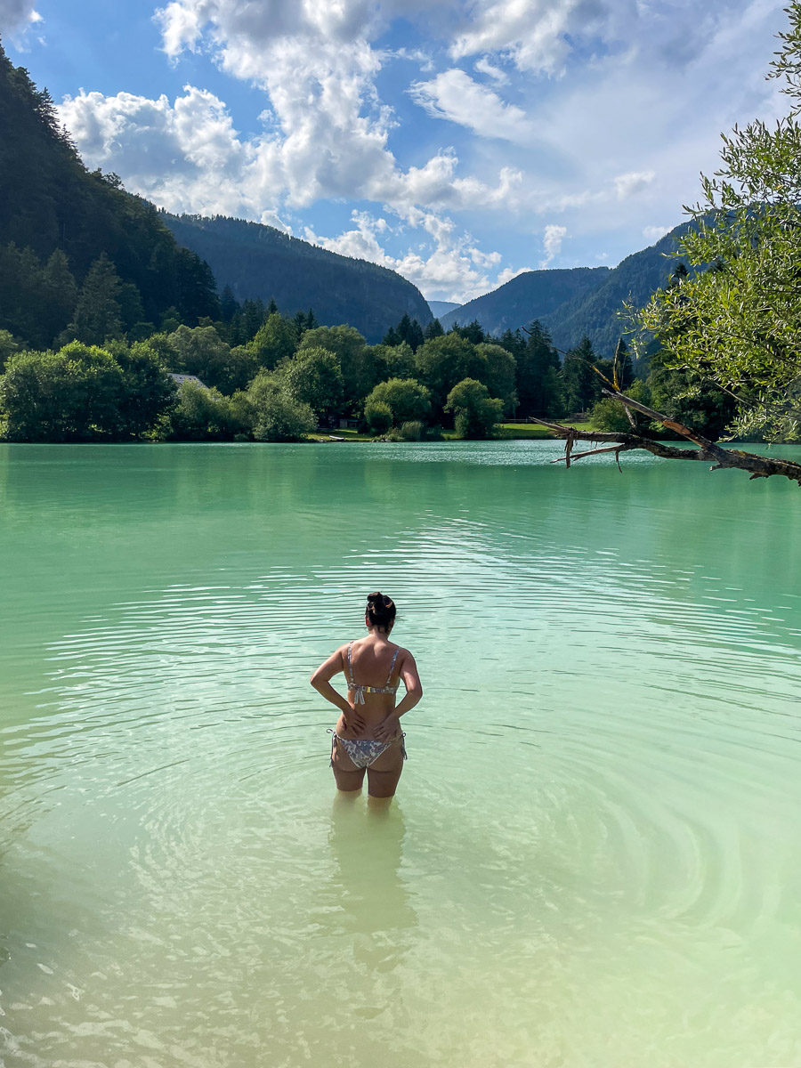 Woman standing in calm green lake with forested mountain valley scenery and blue sky