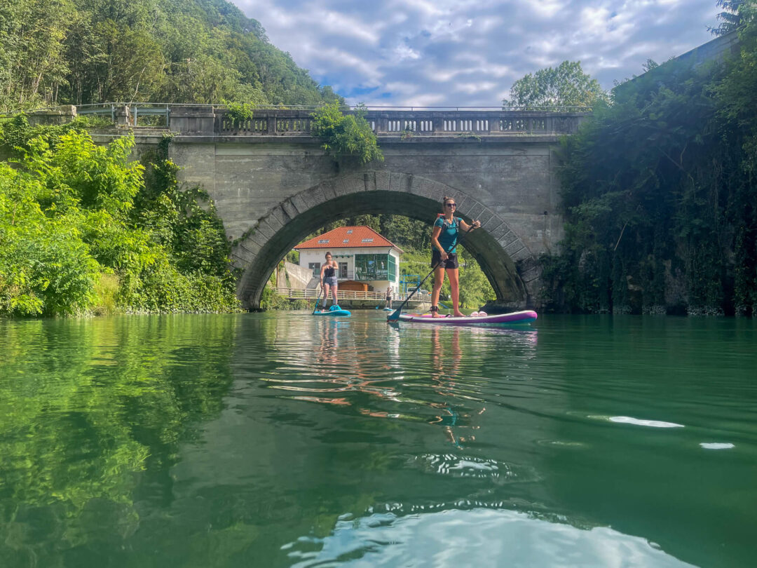 Woman paddleboarding on a calm river under a stone arch bridge, surrounded by lush greenery