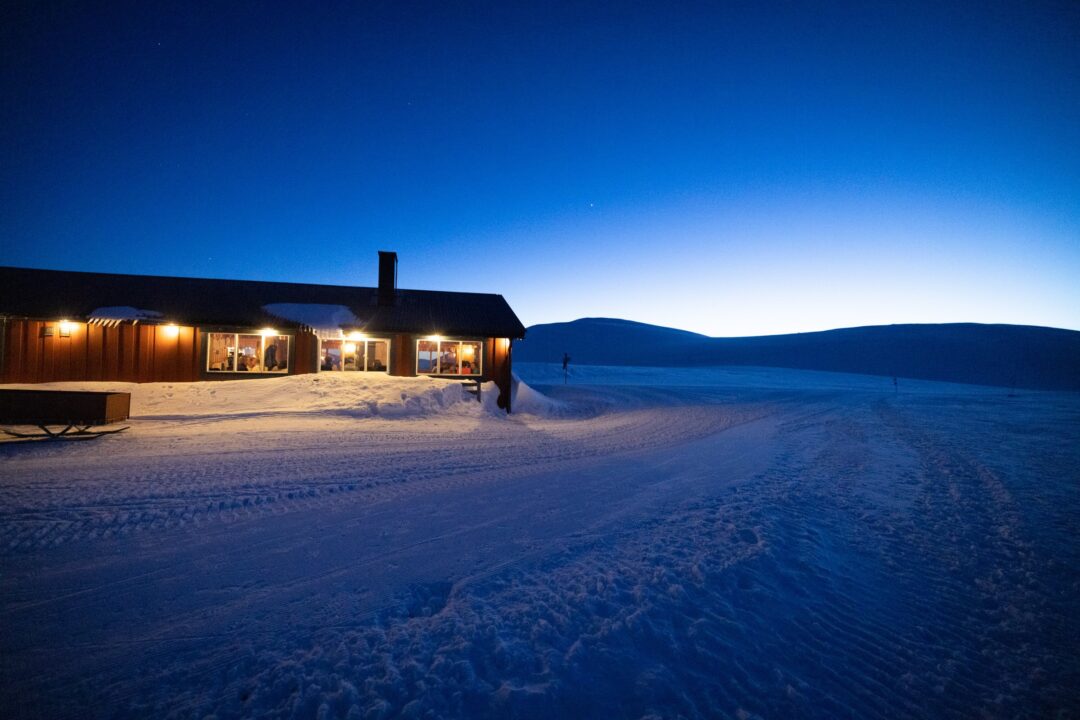 Snow-covered cabin beside a cross-country ski track at dusk in a winter landscape