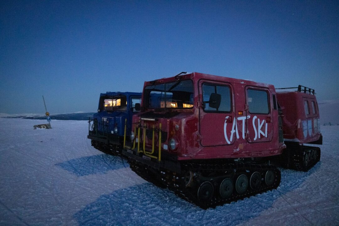 Red tracked snowcat vehicle marked “AT SKI” on snowy terrain at dusk