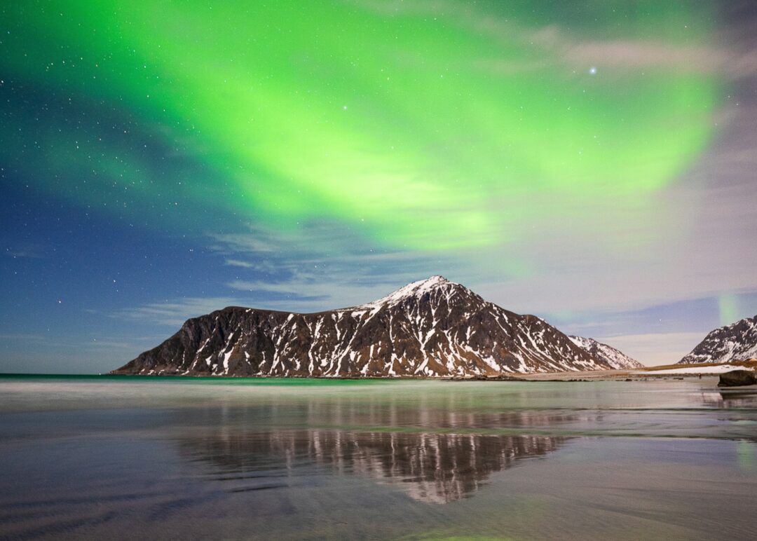 Green northern lights (aurora) over snow-capped mountains and calm shoreline at night