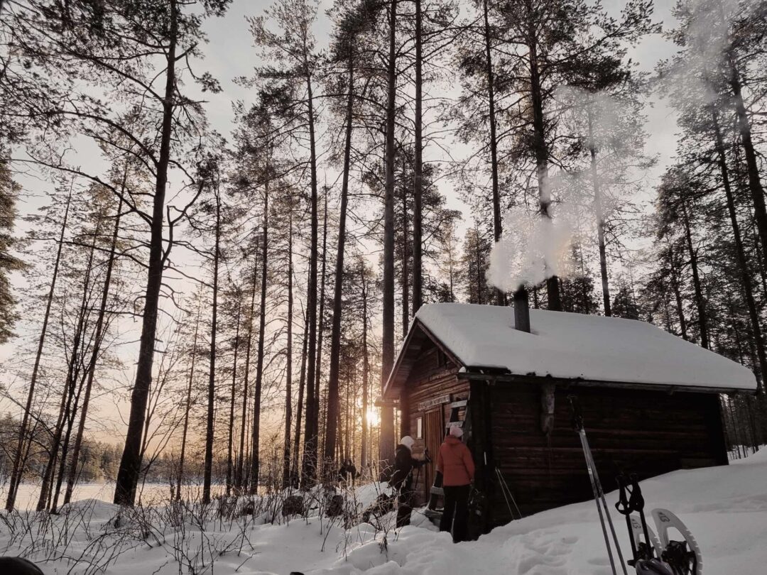 Women walking past a snow-covered log cabin in a pine forest at sunrise