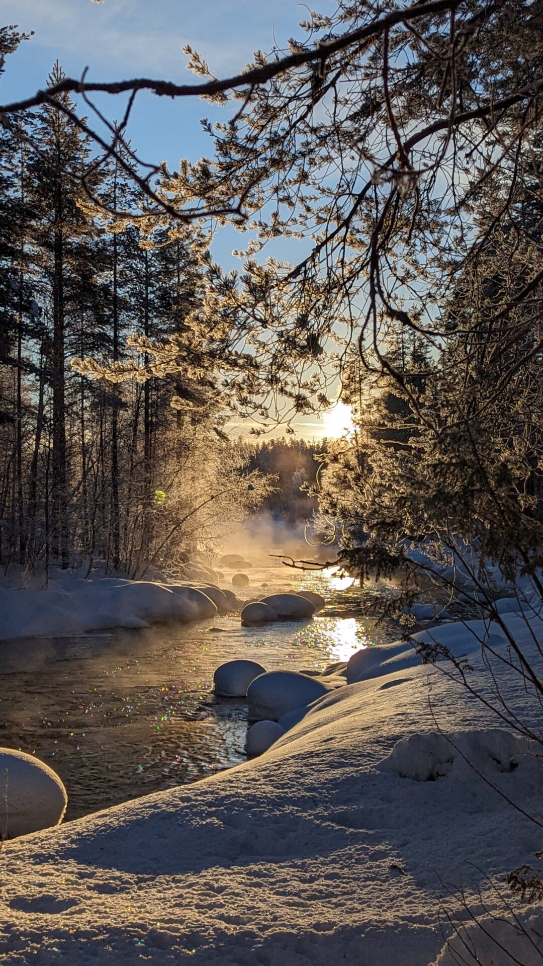 Snow and morning mist over a forest stream at sunrise, with fallen logs and rocks in Finland.