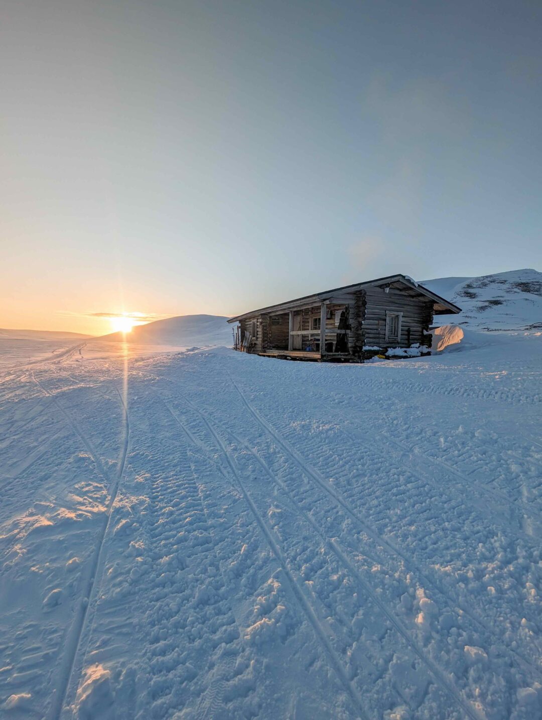 Snowy Arctic piste beside a wooden hut at sunset, with ski tracks in the foreground