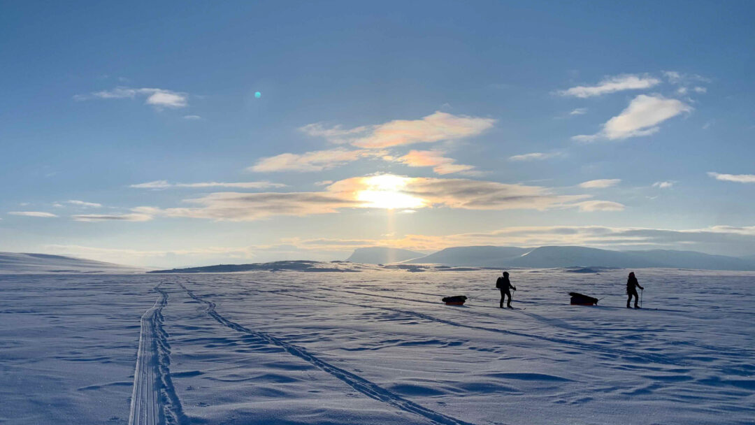 Women cross-country skiing across snowy Arctic landscape at sunset, with tracks across the snow