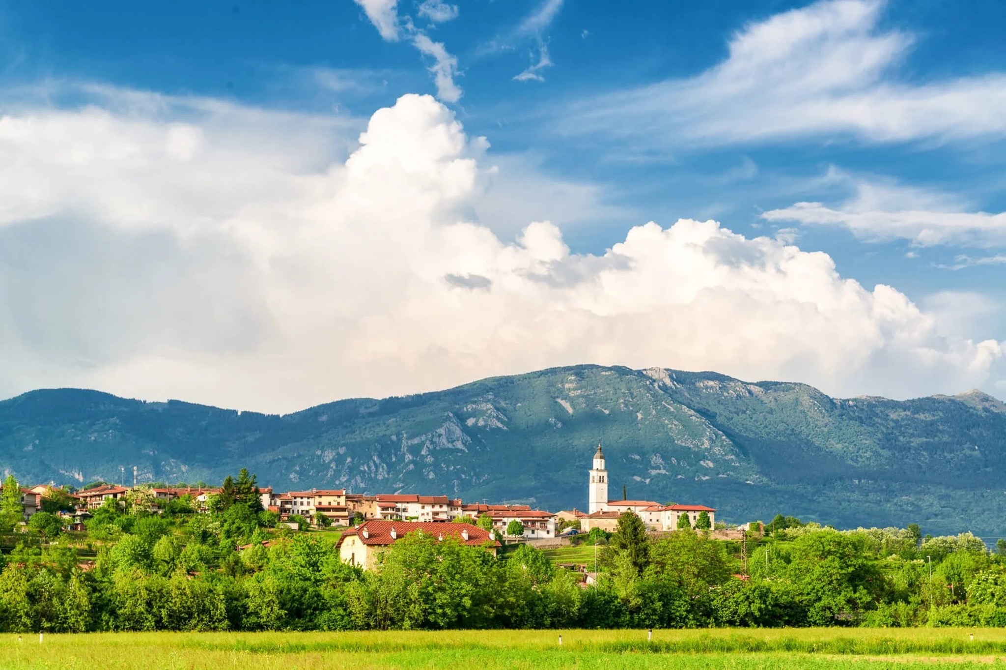 Slovenian hillside village with church tower, surrounded by green fields and mountain peaks under blue sky