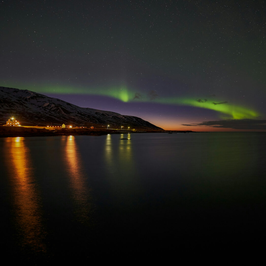 Northern lights over a coastal village and beach at night in Iceland, with green aurora reflecting on the water