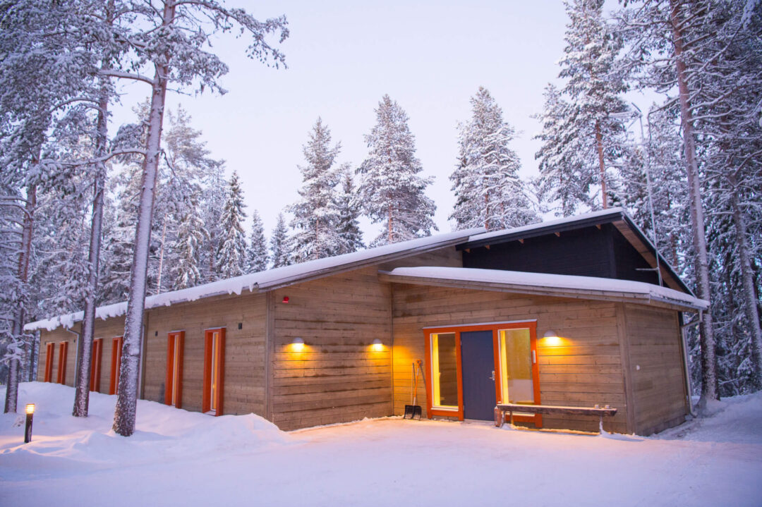 Snow-covered wooden cabin in a pine forest at dusk, with warm lights and an open doorway in Swedish Lapland