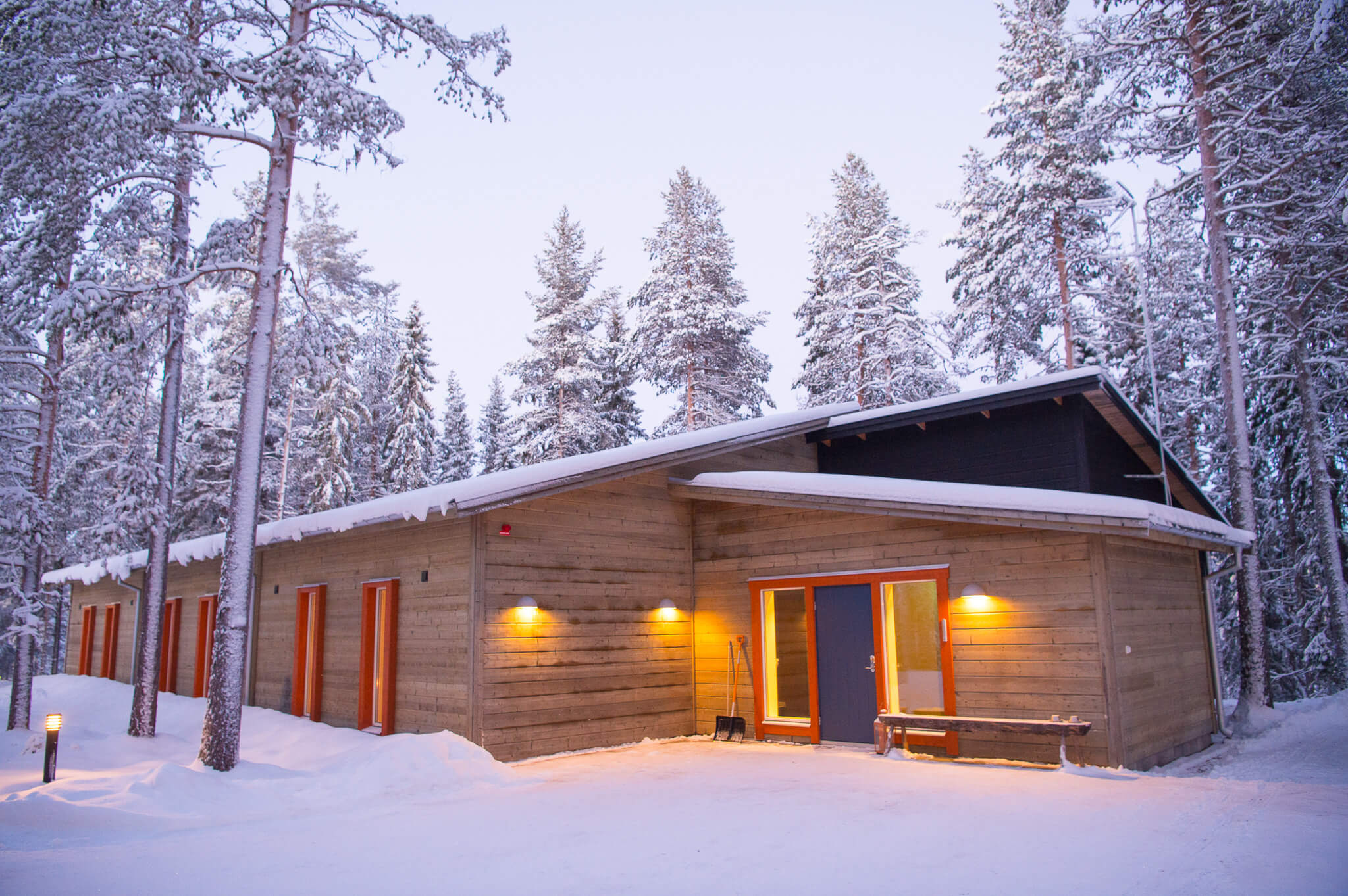 Snow-covered wooden cabin in a pine forest at dusk, with warm lights and an open doorway in Swedish Lapland