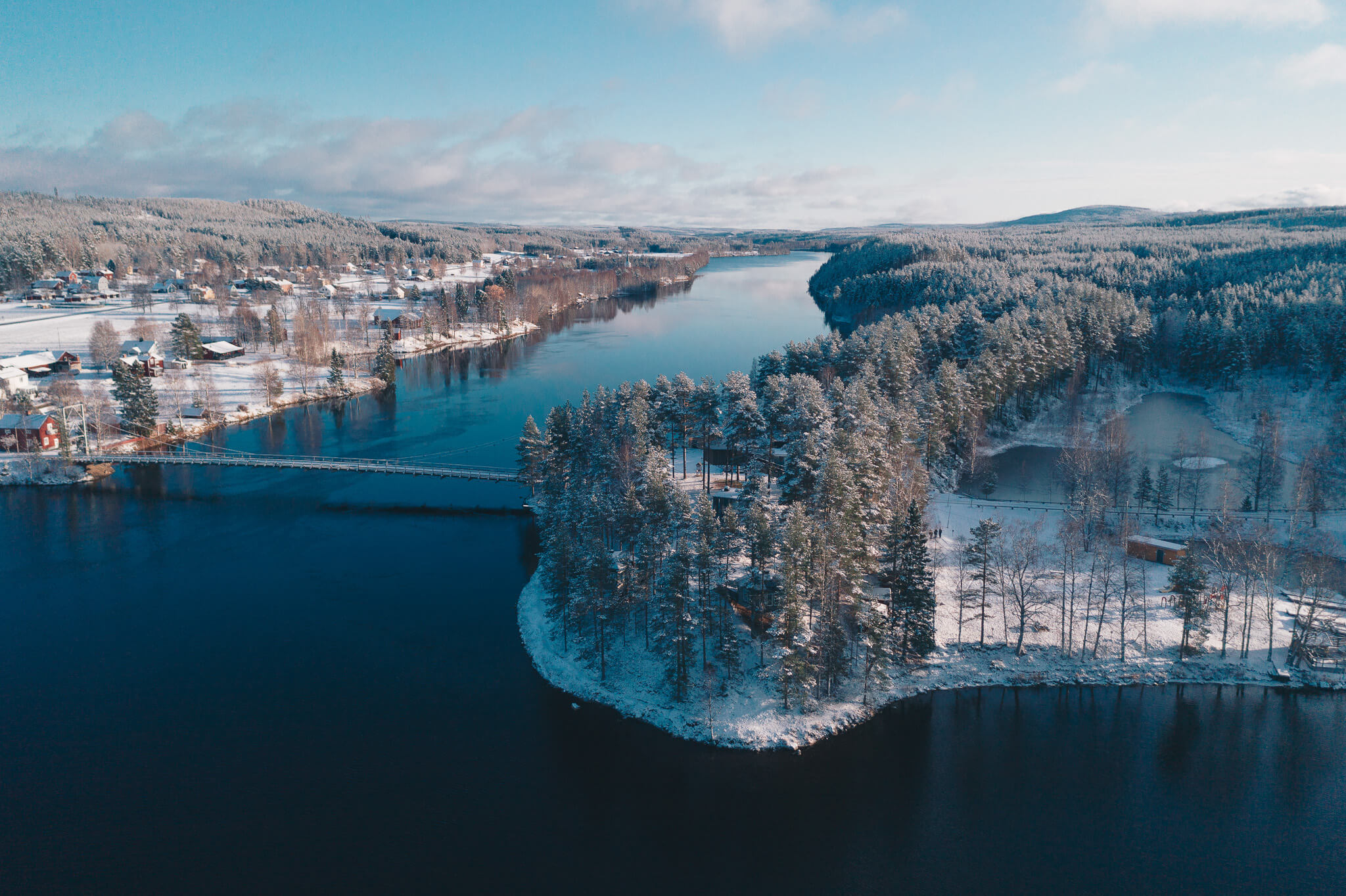 Snowy riverside village and forest along a calm river in Swedish Lapland, aerial view