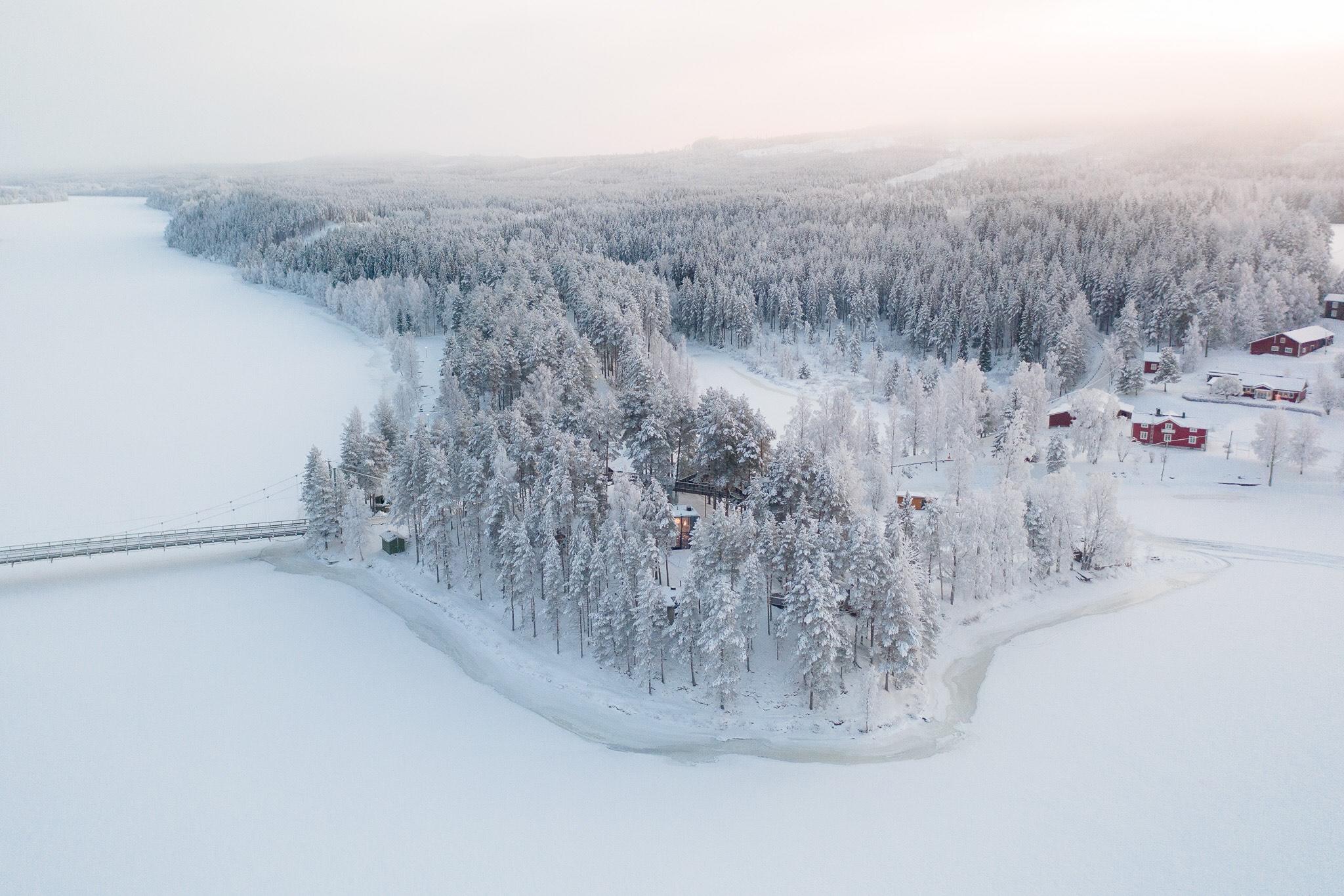 Snow-covered pine forest by a frozen river with small red cabins in snowy Swedish Lapland, aerial view