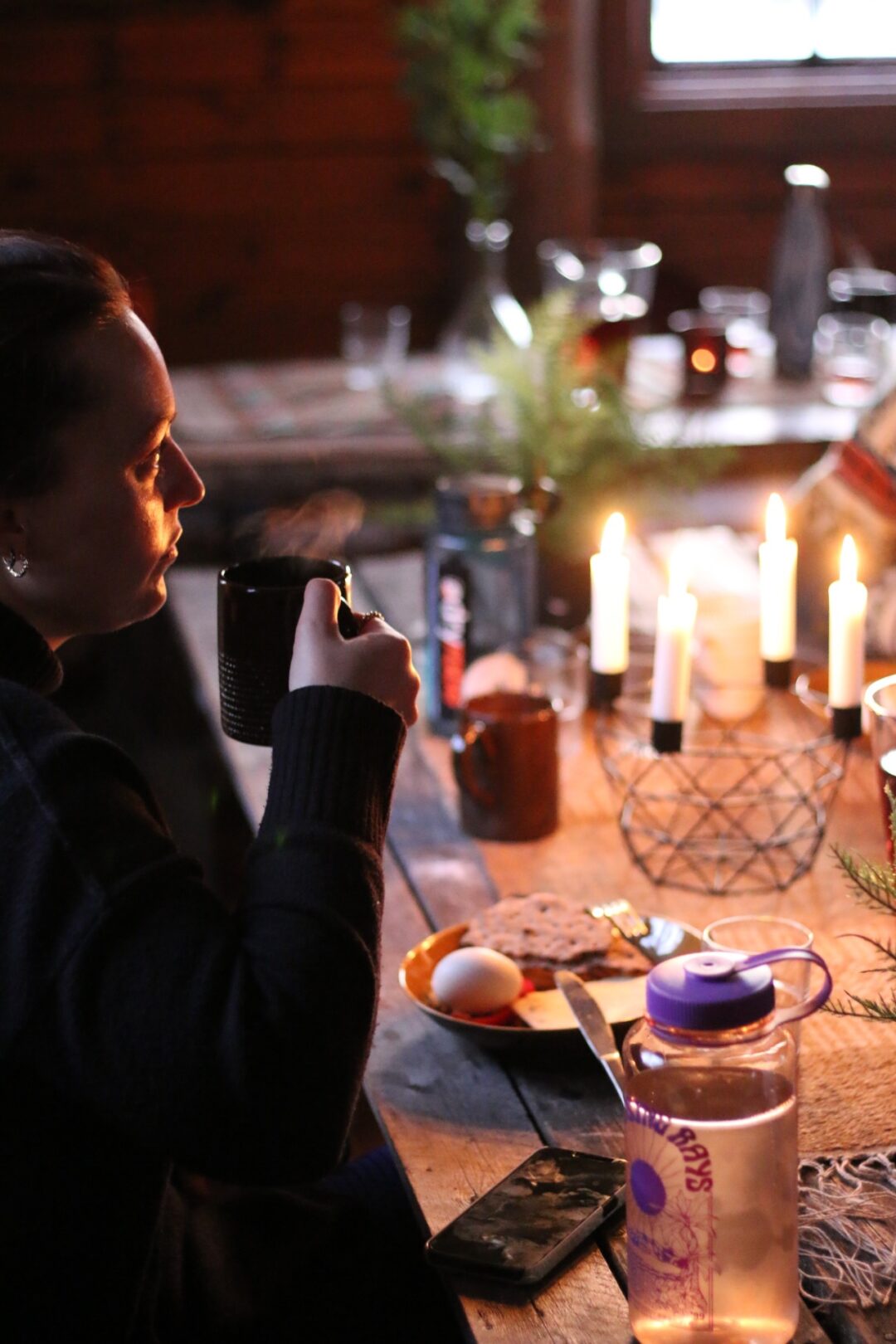 Woman holding a mug by candlelight on a camping table with food and warm drinks indoors